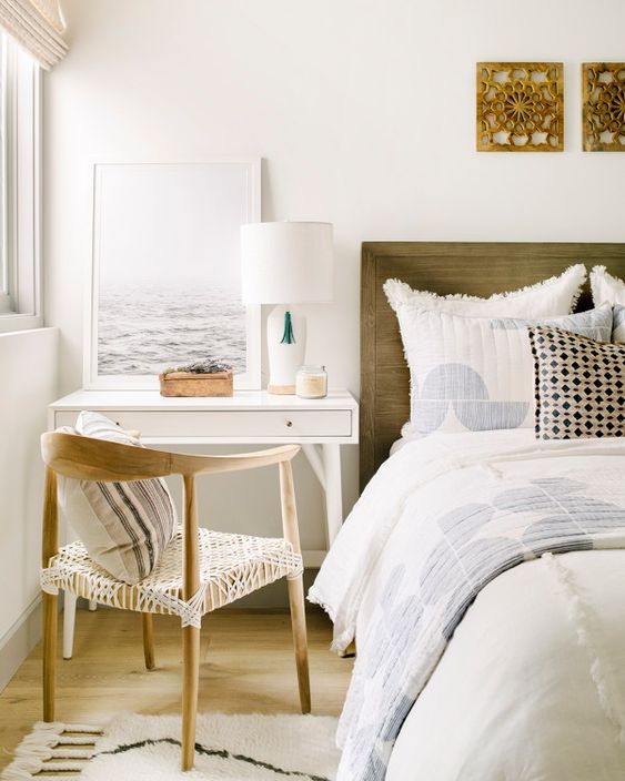 an airy bedroom with a stained bed and neutral bedding, a tiny white desk and a nightstand, some decor and artwork