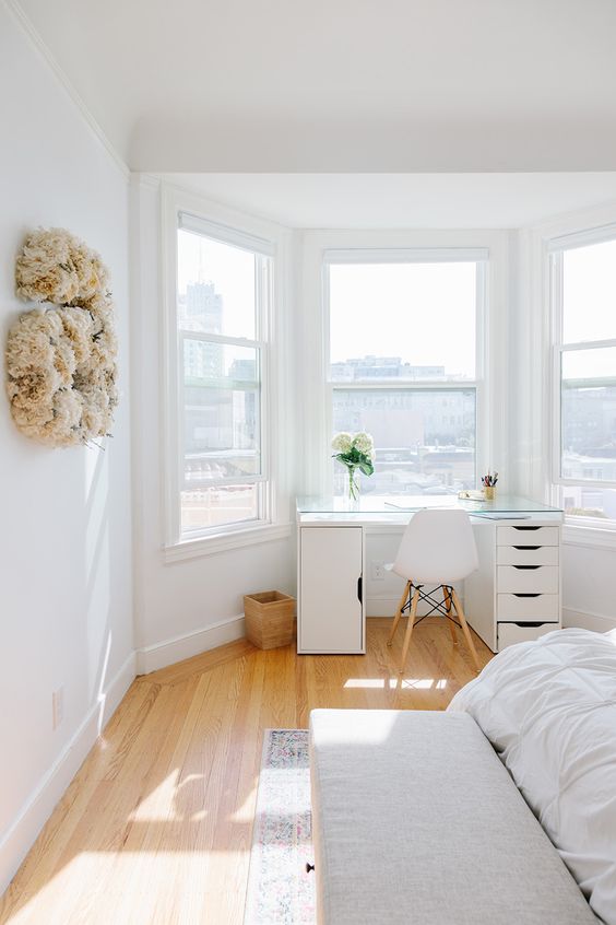 an airy bedroom with a white bed and bedding, a bench, a white desk in the window alcove and some decor