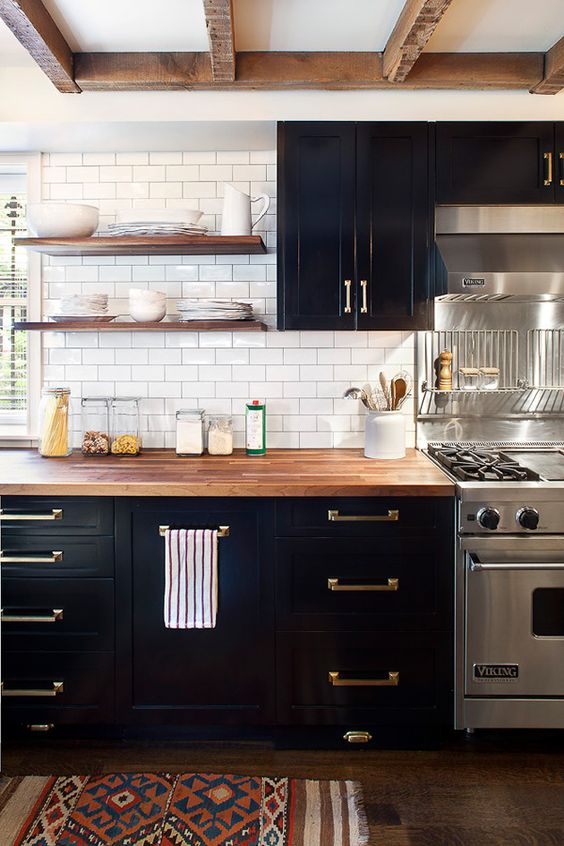 a classy black farmhouse kitchen with a white subway tile backsplash, butcherblock countertops and stained shelves plus brass handles is pure chic