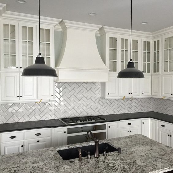 a classic white kitchen with a large statement hood, subway tile laid with a diagonal herringbone pattern, black countertops and pendant lamps