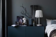 a moody and cool bedroom with grey walls, a teal cabinet, a bed with neutral bedding, some decor and a table lamp is a chic and lovely space