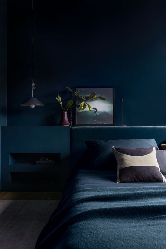 a beautiful dark bedroom with navy walls and a built-in matching niche, a navy upholstered bed with navy bedding, some art and a pendant lamp