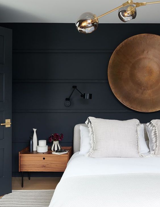 a beautiful moody bedroom with soot walls and a door, a grey upholstered bed with neutral bedding, stained nightstands, some lamps and a wooden bowl on the wall