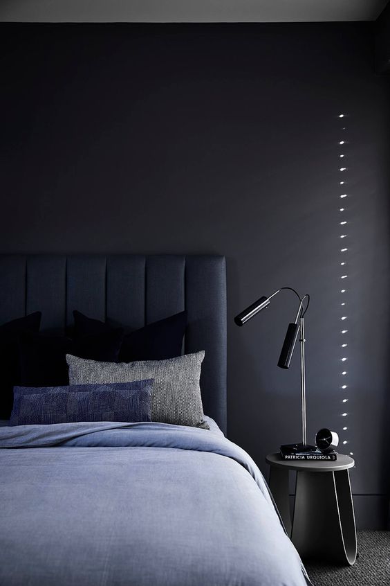a chic and peaceful moody bedroom with a black accent wall, a navy upholstered bed and blue bedding, a nightstand, black table lamps