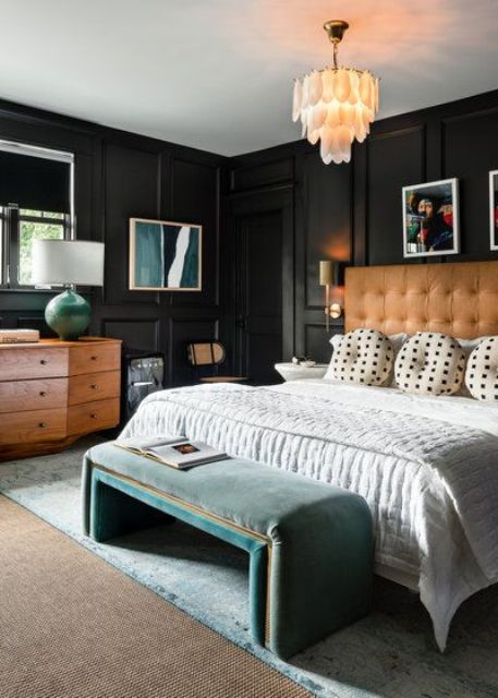 a chic moody bedroom with black molded walls, an amber leather bed with neutral bedding, a green upholstered bench and a stained dresser