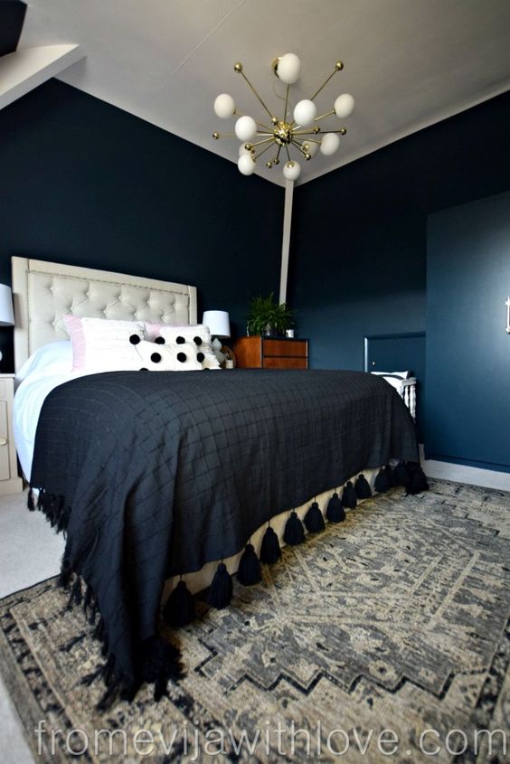 a dark bedroom with navy walls, some niches, an upholstered bed with contrasting bedding, a printed rug and a cool chandelier