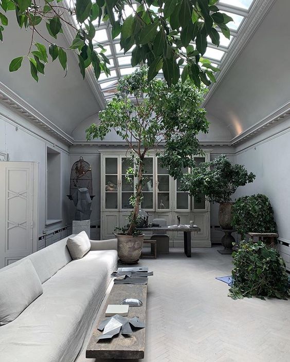 a grey living room with a large skylight, a storage unit, a low neutral sofa, a stained bench, a working space and potted greenery