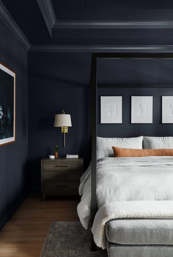 a midnight blue bedroom with a black canopy bed with neutral bedding, a neutral upholstered bench, a dark nightstand, some artwork