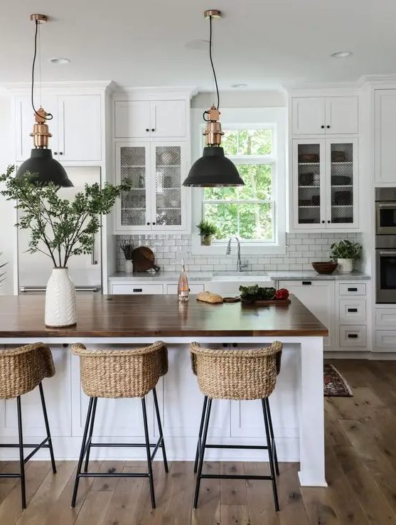 a modern country kitchen in white, with cool cabinets, a large kitchen island with a butcherblock countertop, pendant lamps and woven stools