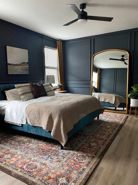 a moody and peaceful bedroom with soot walls, a blue upholstered bed with neutral bedding, a large arched floor mirror and a printed rug