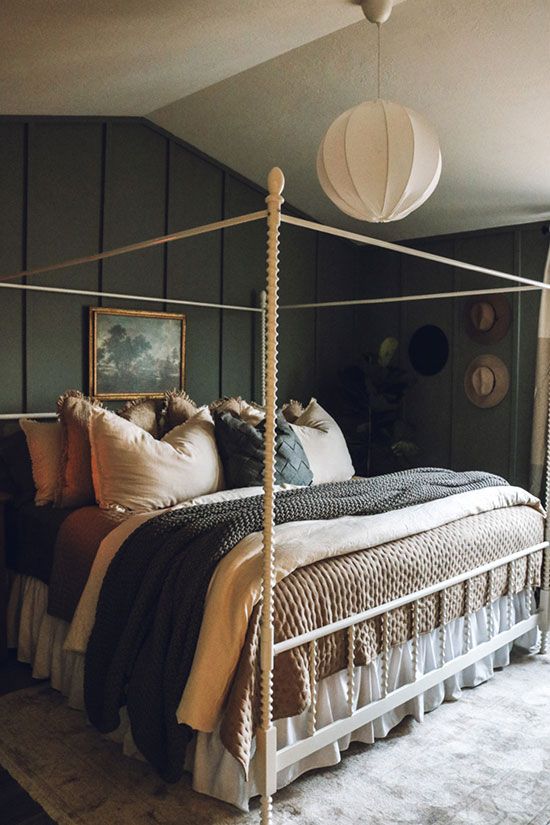 a moody attic bedroom with graphite grey walls, a white canopy bed with lots of neutral and dark pillows, a white pendant lamp and a hat gallery wall