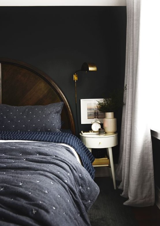 a moody bedorom with dark walls, a dark wooden bed, navy and blue bedding, a round nightstand and some greenery