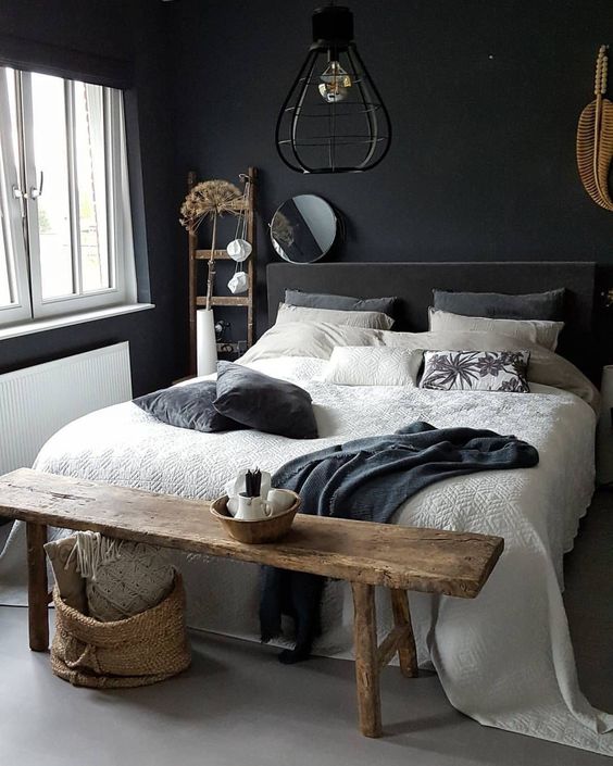 a moody bedroom with black walls, a grey upholstered bed, neutral bedding, a rough stained bench, baskets and a ladder plus some wabi-sabi decor