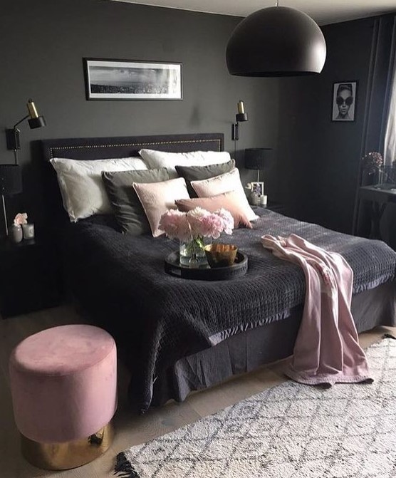 a moody bedroom with grey walls, a black bed, nightstand and a pendant lamp, pink and black bedding and a pink ottoman