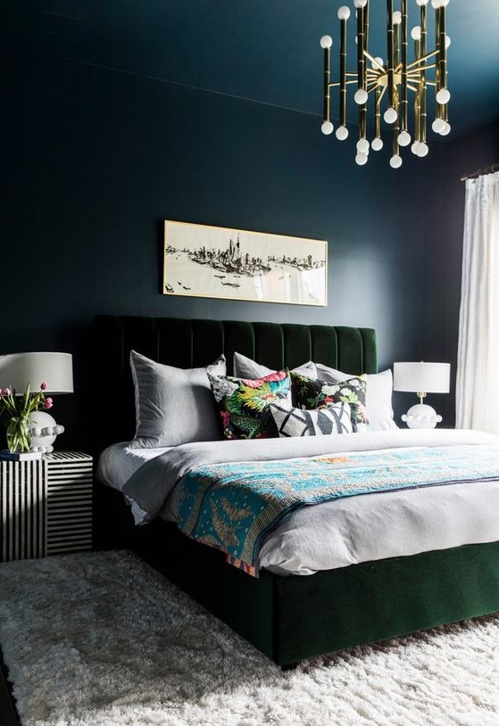 a moody bedroom with navy walls and a ceiling, a dark green velvet bed, a glam gold chandelier and striped nightstands