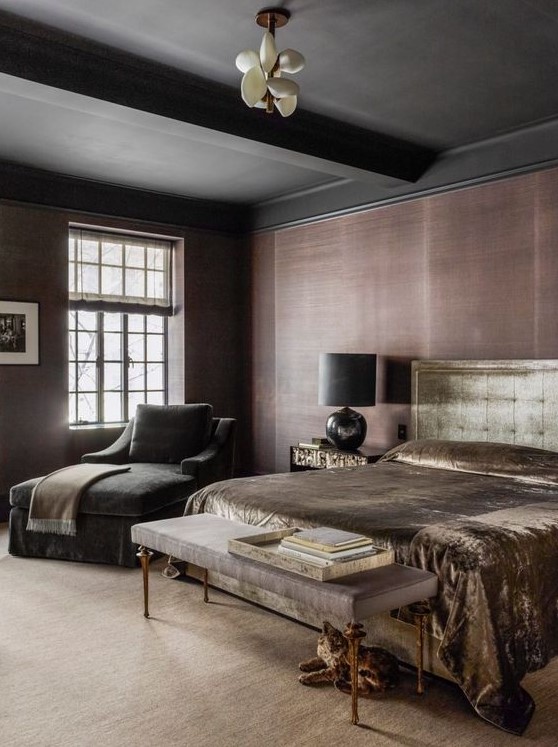 a moody bedroom with taupe grasscloth wallpaper, a black ceiling and beams, a gold velvet bed, a taupe velvet bench and a black lounger