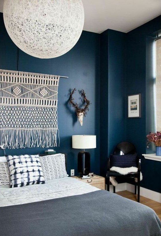a moody boho bedroom with navy walls, black chairs, a macrame hanging, a woven lamp and printed bedding
