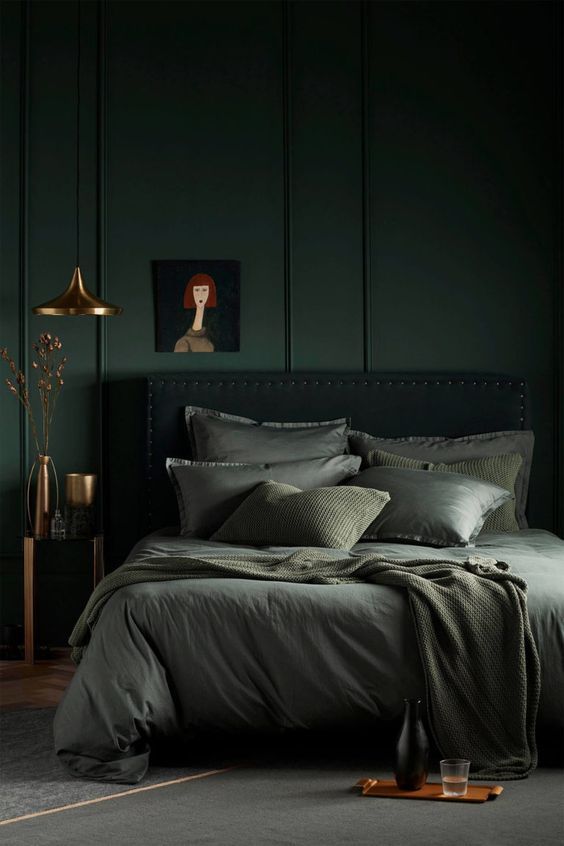 a moody dark green bedroom with a black upholstered bed, green bedding, a nightstand with gold vases and an artwork