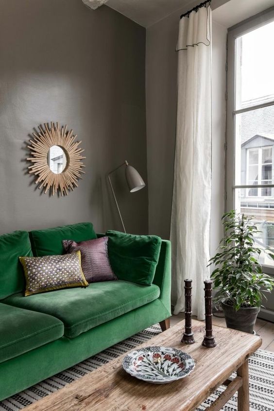 a moody living room with grey walls, a bold grene sofa, a stained bench as a coffee table, a burst mirror and a taupe floor lamp