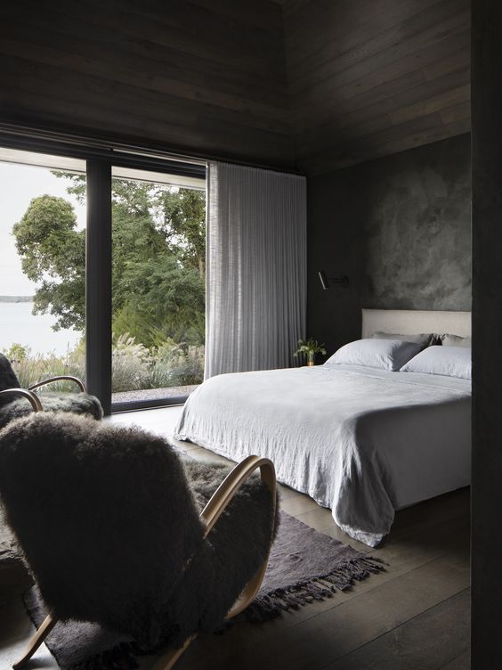 a pretty moody bedroom with stained wood on the ceiling, grey plaster walls, a neutral upholstered bed with neutral bedding, faux fur chairs and a glazed wall