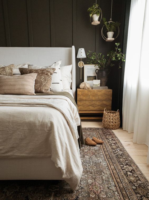 a welcoming moody bedroom with a soot accent wall, a neutral upholstered bed with neutral bedding, a printed rug, pendant plants and an inlay nightstand