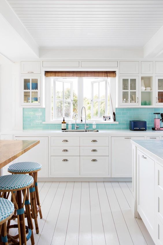 a white coastal kitchen with shaker cabinets, a turquoise subway tile backsplash, a stained table and blue stools