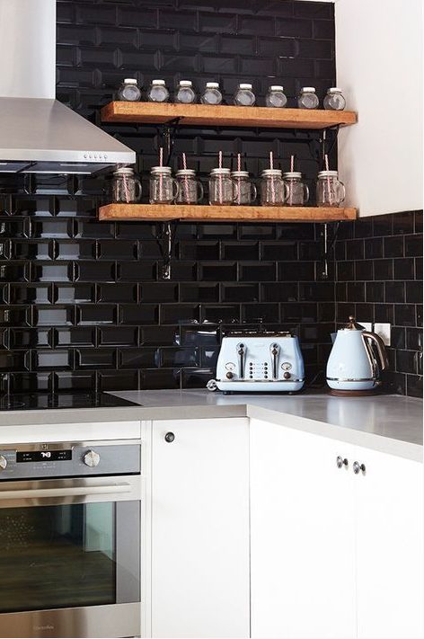 a white kitchen with grey countertops and a black glossy tile backsplash that takes two walls plus floating stained shelves for spices
