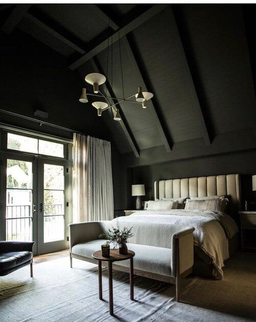 an attic bedroom with black walls and a ceiling, a neutral upholstered bed with neutral bedding, a neutral bench, a black chair and a chandelier