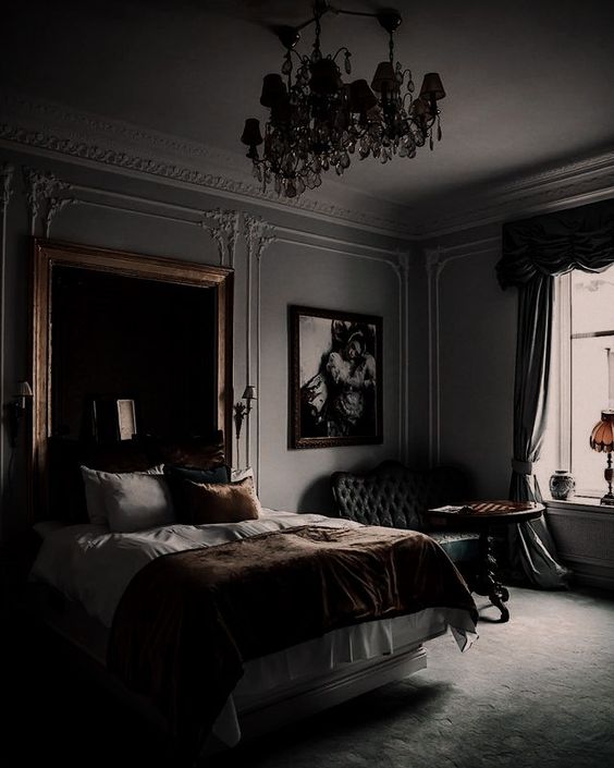 an exquisite moody bedroom with grey walls, a bed with neutral bedding, an oversized wall mirror, a refined black sofa and chic chandeliers