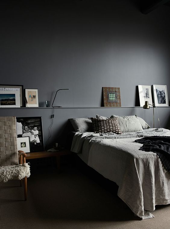 a moody bedroom with graphite grey walls, a bed with greyish bedding, a ledge with artwork and a woven chair plus some pillows