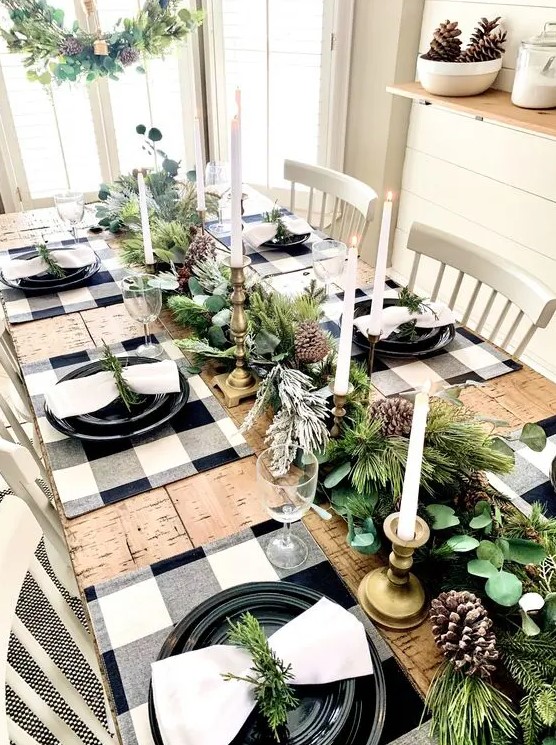 a farmhouse Christmas table with an evergreen and pinecone runner, tall and thin candles, buffalo check placemats, black plates