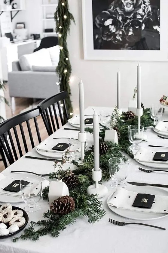 a minimalist Christmas tablescape with white linens, grey candles in white candleholders, an evergreen runner with pinecones and little houses