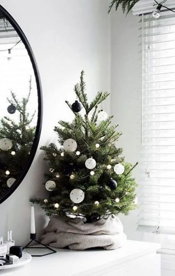 a modern Christmas tree decorated with blak and white ornaments and pompom lights plus an old sweater as a tree skirt