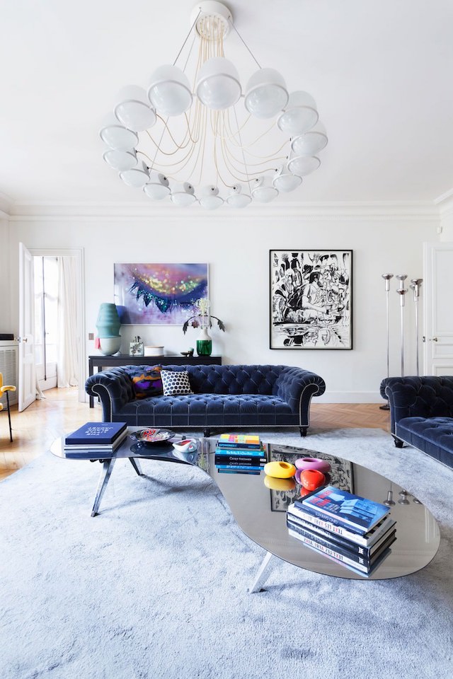The living room is dotted with colors   navy velvet furniture and some bold artworks and a mirror kidney shaped table catches an eye