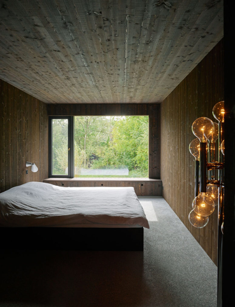 This is a master bedroom, fully clad with dark rough wood, there's a glass wall with a storage underneath and some industrial light fixtures