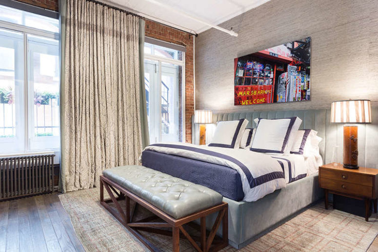 the bedroom is decorated in muted shades and neutrals, there's a grey upholstered bed and bench, an exposed brick wall and a bold photo