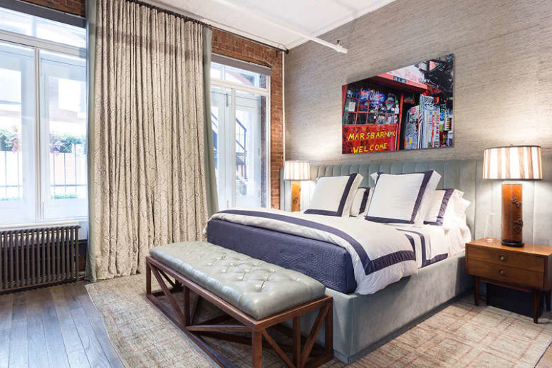 the bedroom is decorated in muted shades and neutrals, there's a grey upholstered bed and bench, an exposed brick wall and a bold photo the bedroom is decorated in muted shades and neutrals, there's a grey upholstered bed and bench, an exposed brick wall and a bold photo