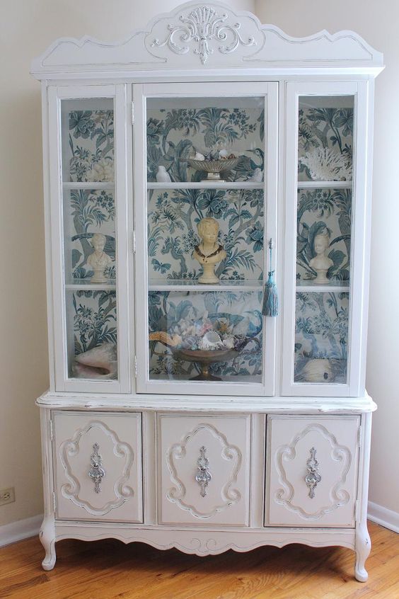 a white vintage cabinet with blue and white wallpaper inside for displaying souvenirs