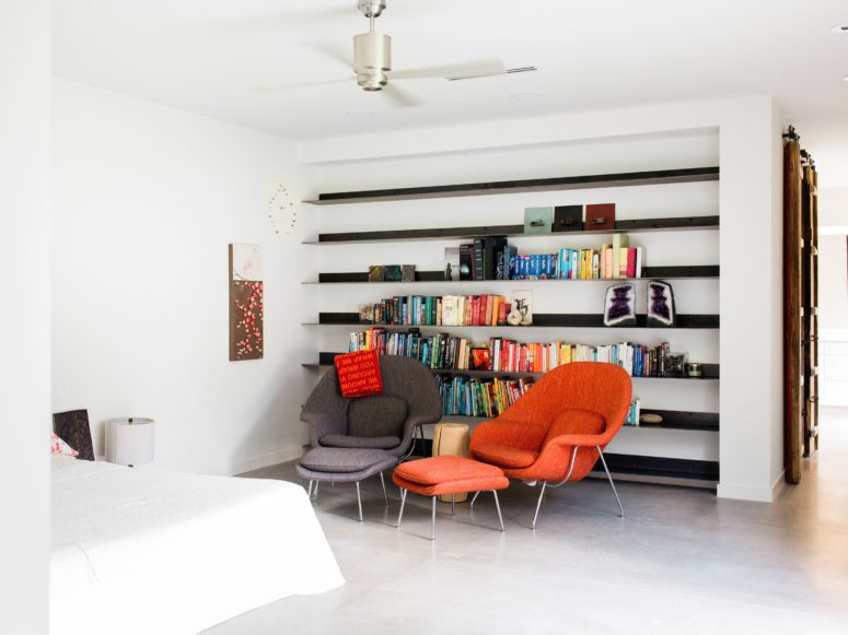 The bedroom features a large bed and a sitting zone with colorful chairs and one wall is taken by open shelves