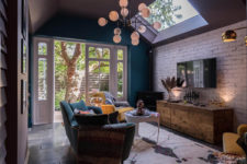 The living room features a teal wall and velvet sofa in the same color, some jewel toned chairs, skylights for additional natural light