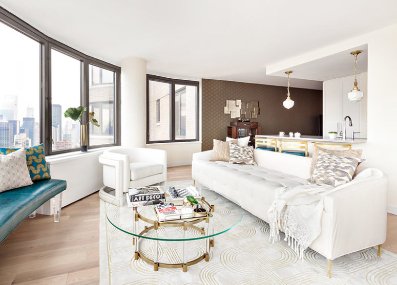 The large rounded room done in neutral shades is spruced up with a teal leather bench, glass, brass and acrylic touches