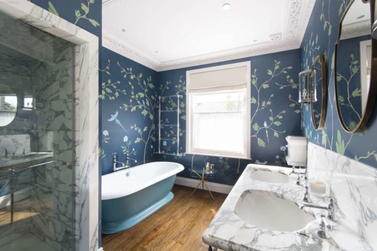 The master bathroom is done with navy floral print walls, a marble vanity and shower and a vintage blue bathtub