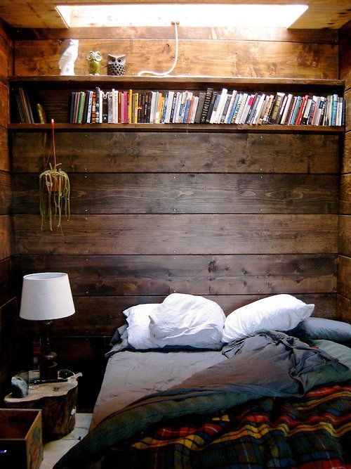 a cozy small rustic bedroom with a stained wooden wall and a matching bookshelf over the bed
