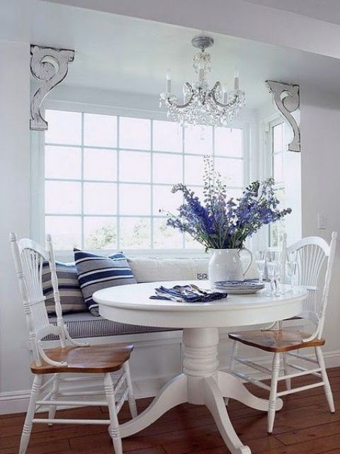 a coastal breakfast nook with an upholstered windowsill bench, a round pedestal table and some chairs