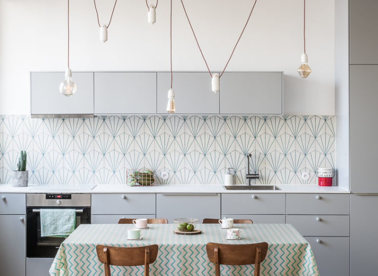 The geometric kitchen backsplash stands out nextot the grey kitchen cabinets, and lamps hanging on red cord add an industrial feel