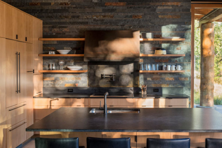 The kitchen is done with light-colored wooden cabinets, darkened metal appliances and stone countertops, the walls are clad with reclaimed wood