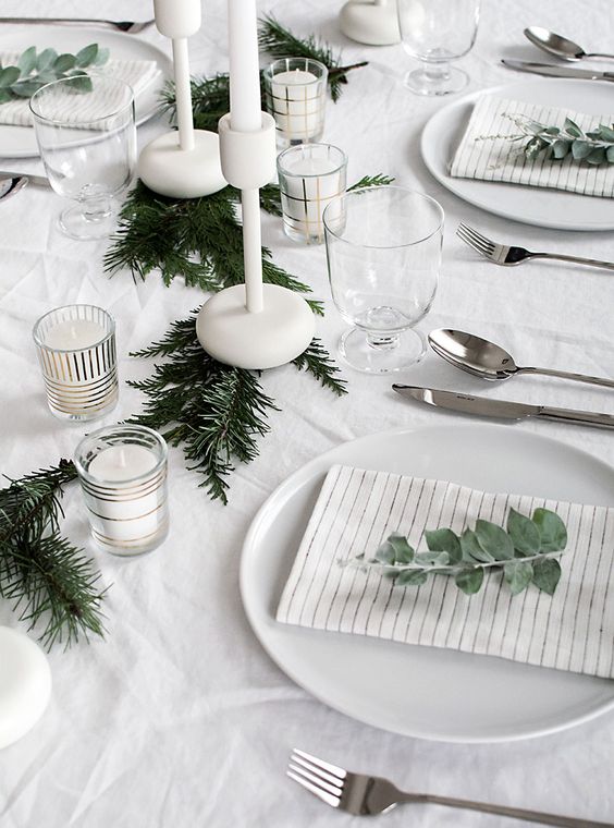 a minimalist table setting with an evergreen garland, white candles and eucalyptus for marking each place setting