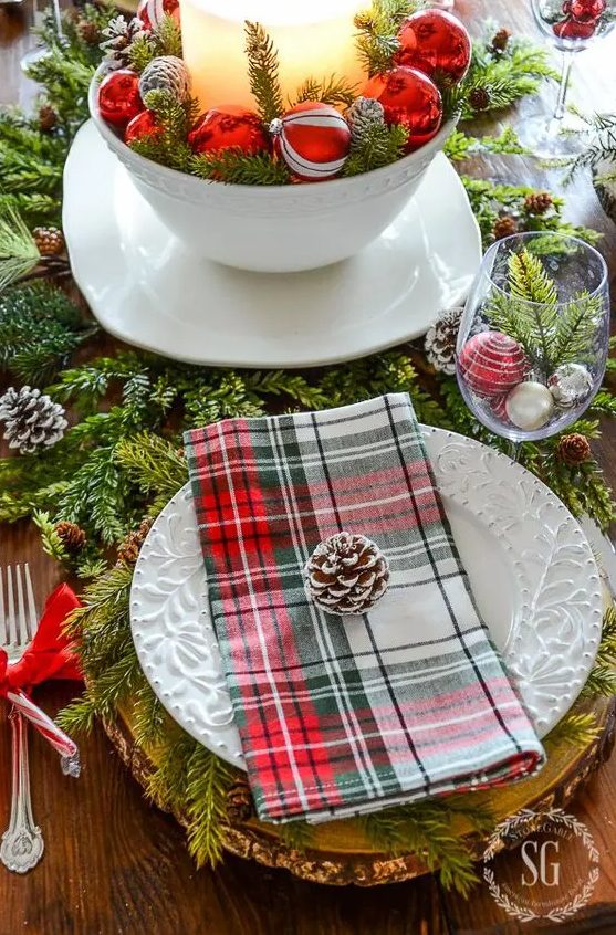 a cozy traditional table setting with evergreens, pinecones, plaid napkins, a tree slice as a placemat and ornaments