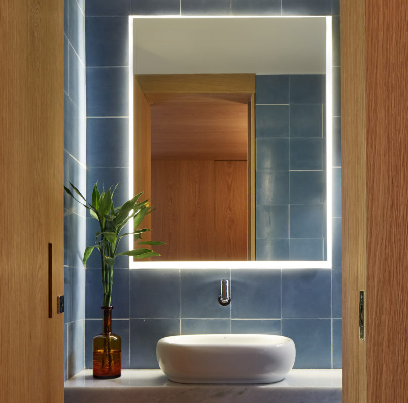 The powder room is done with blue tiles, a marble countertop and a sculptural sink and a lit up mirror