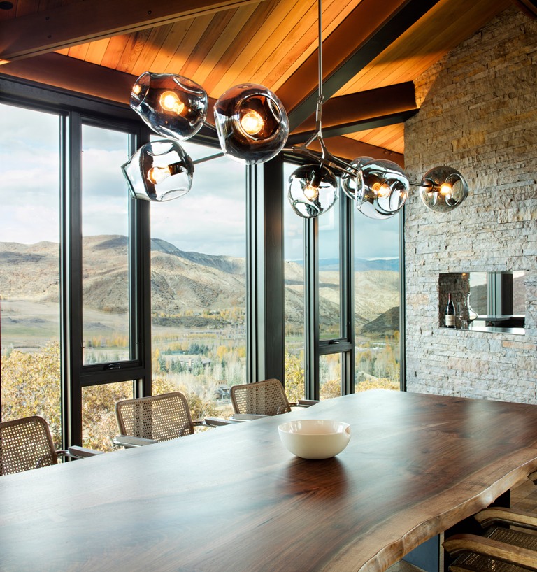 The inner dining room is done with a natural stone wall, extensive glazing, a wooden dining table with a liveedge and a catchy chandelier